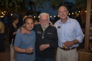 Three men are smiling outside while holding beers.