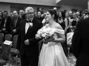 A man in a tuxedo is walking next to his daughter. She is wearing a white wedding gown and holding a bouquet of flowers. Her arm is linked in his.