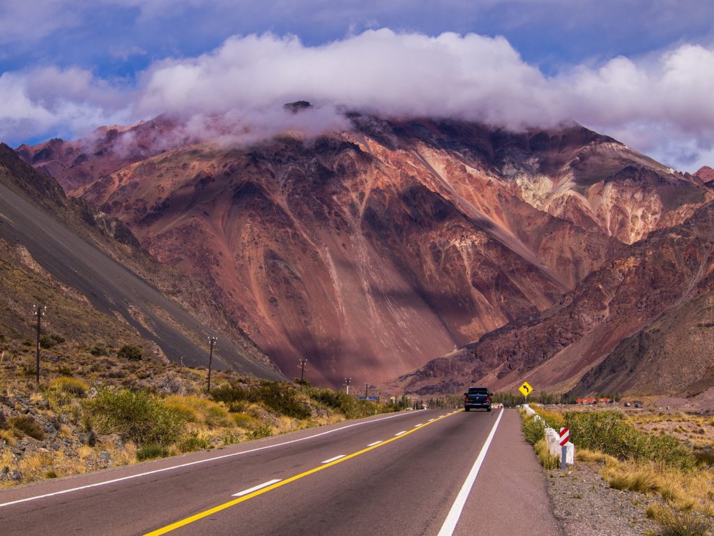 Dark pickup truck driving on a two-lane highway leading into mountains topped with fluffy white cloud.