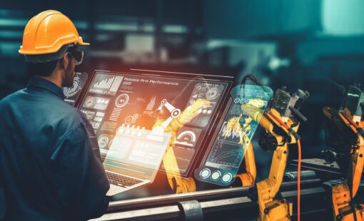 Engineer in a yellow hard hat overseeing two yellow smart industry robot arms and a large monitor for digital factory.