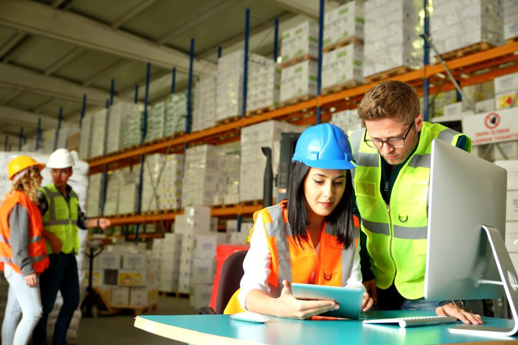 Men and women wearing neon protective vests looking at a tablet device inside a warehouse with two other workers in hardhats in the background