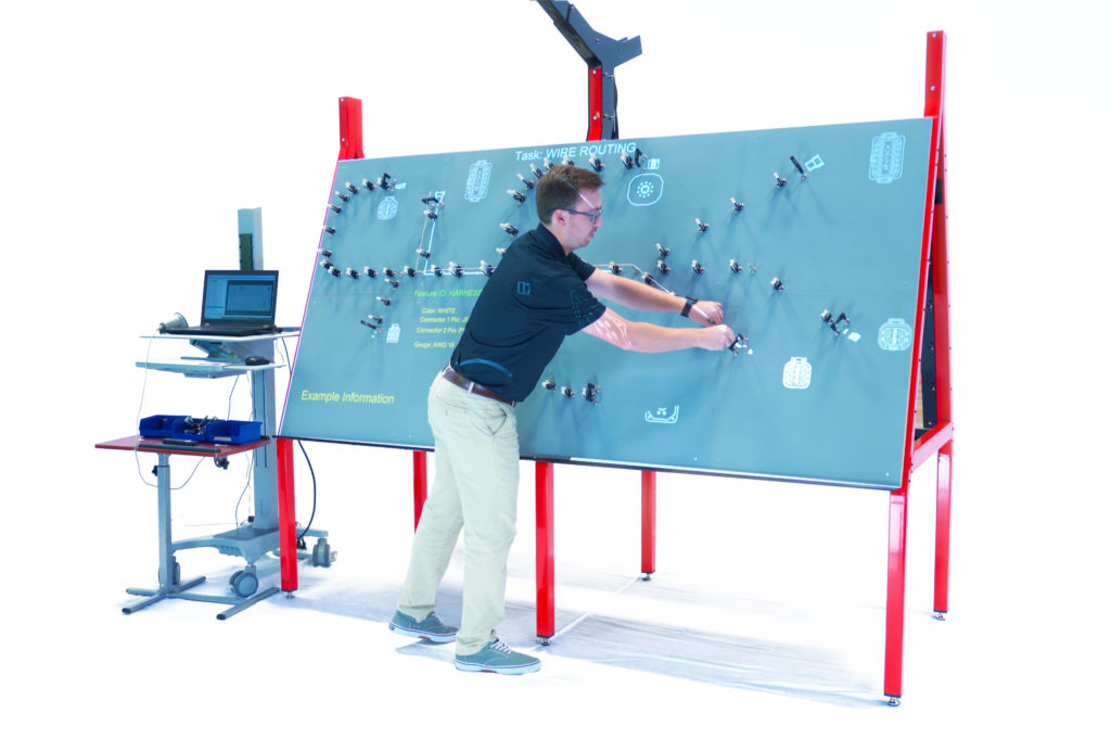 Man in a black shirt is placing connectors on a large magnetic wire harness formboard. Placement diagrams are projected onto the board.