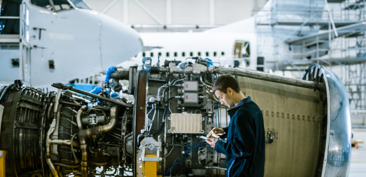 An aircraft maintenance technician inspects a jet engine inside a hangar while holding a tablet, with a commercial airplane in the background.