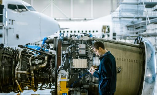 An aircraft maintenance technician inspects a jet engine inside a hangar while holding a tablet, with a commercial airplane in the background.