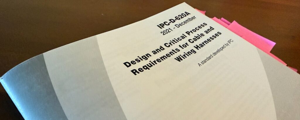 Photograph of the IPC-D-620A: Design and Critical Process Requirements for Cable and Wiring Harnesses standard, 2021 edition, with multiple pink sticky notes indicating referenced sections.