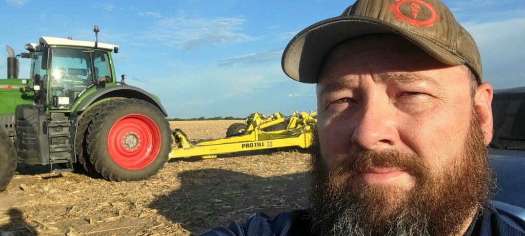 Mike Bayliff standing outdoors in front of a yellow agricultural windrower wearing a hat.