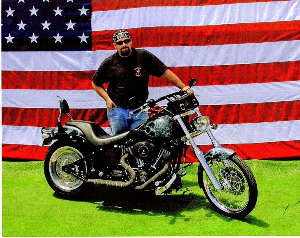 Man standing next to a motorcycle in front of an American flag, wearing a black shirt and a bandana on his head.